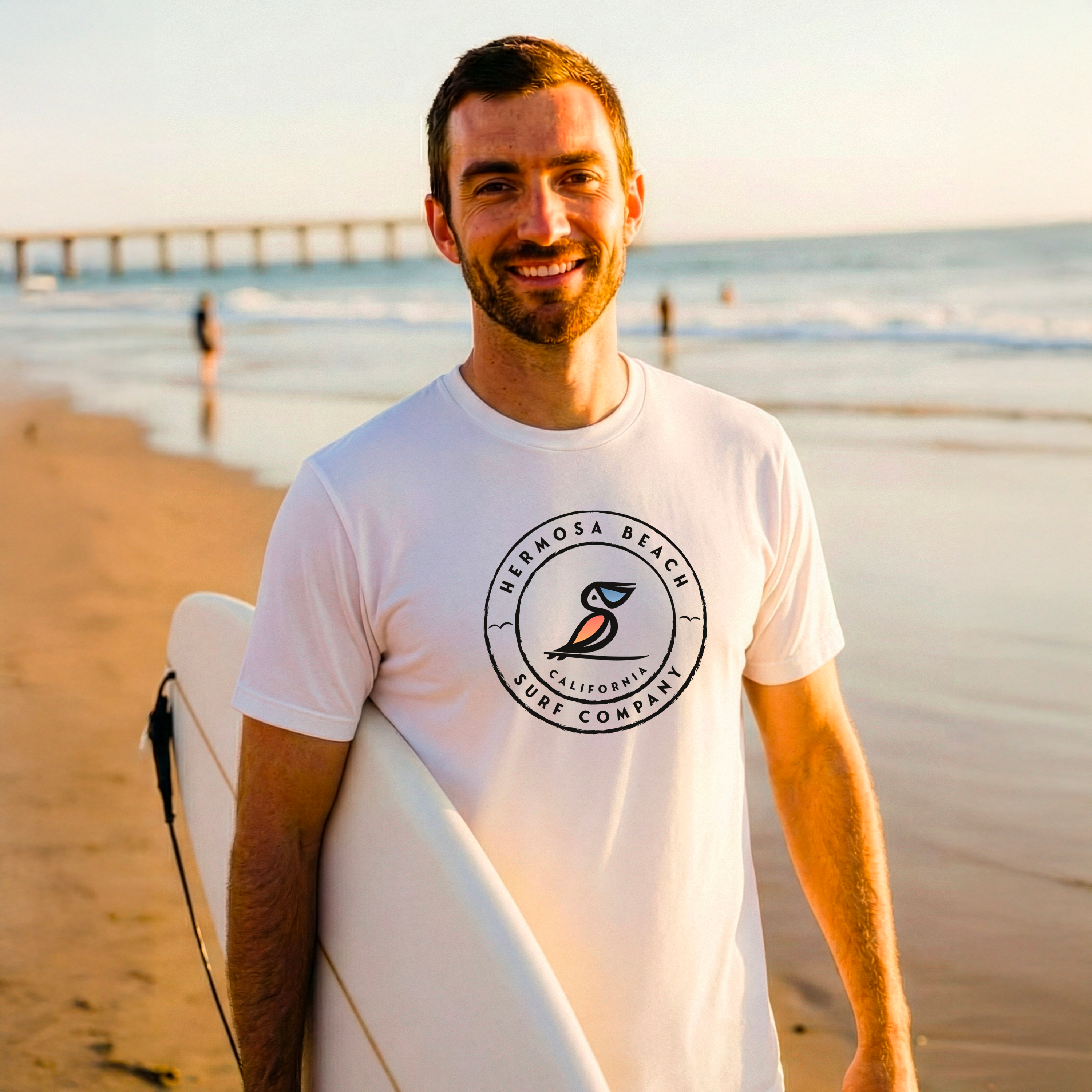 Man holding a surfboard on a beach wearing a Hermosa Beach Surf Company t-shirt.