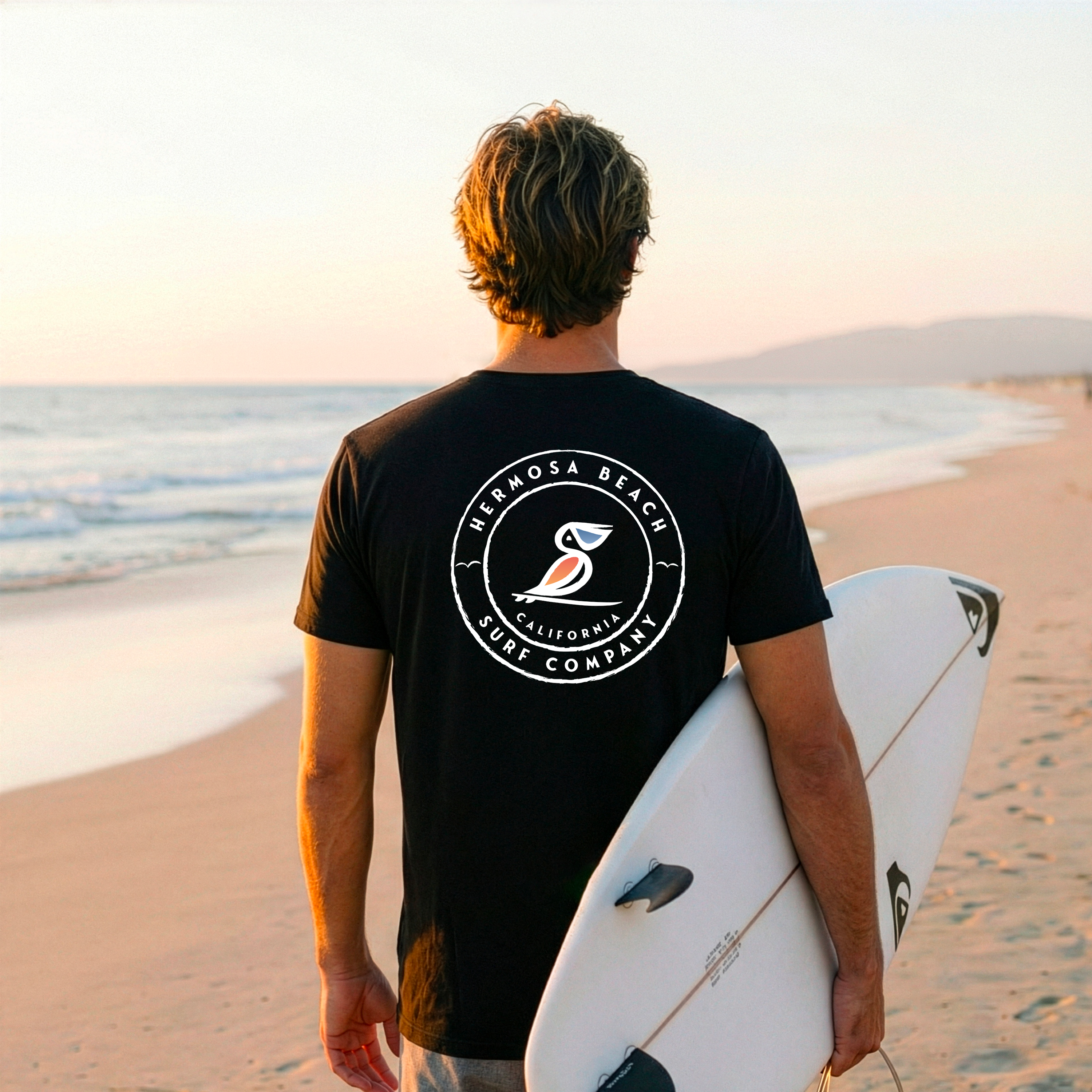 Person holding a surfboard on a beach with a sunset, wearing a black t-shirt with a logo.