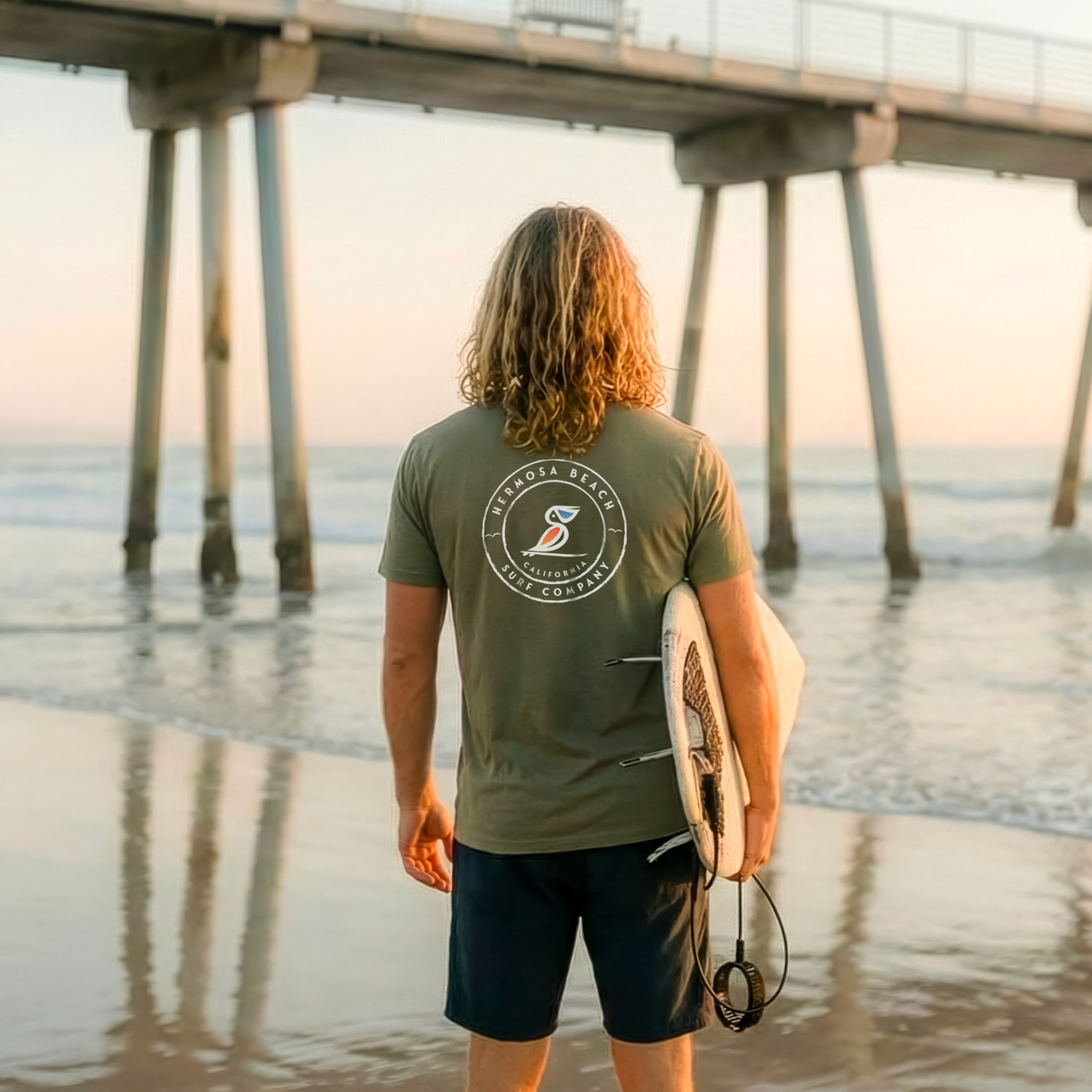 Man holding a surfboard on a beach with a pier in the background