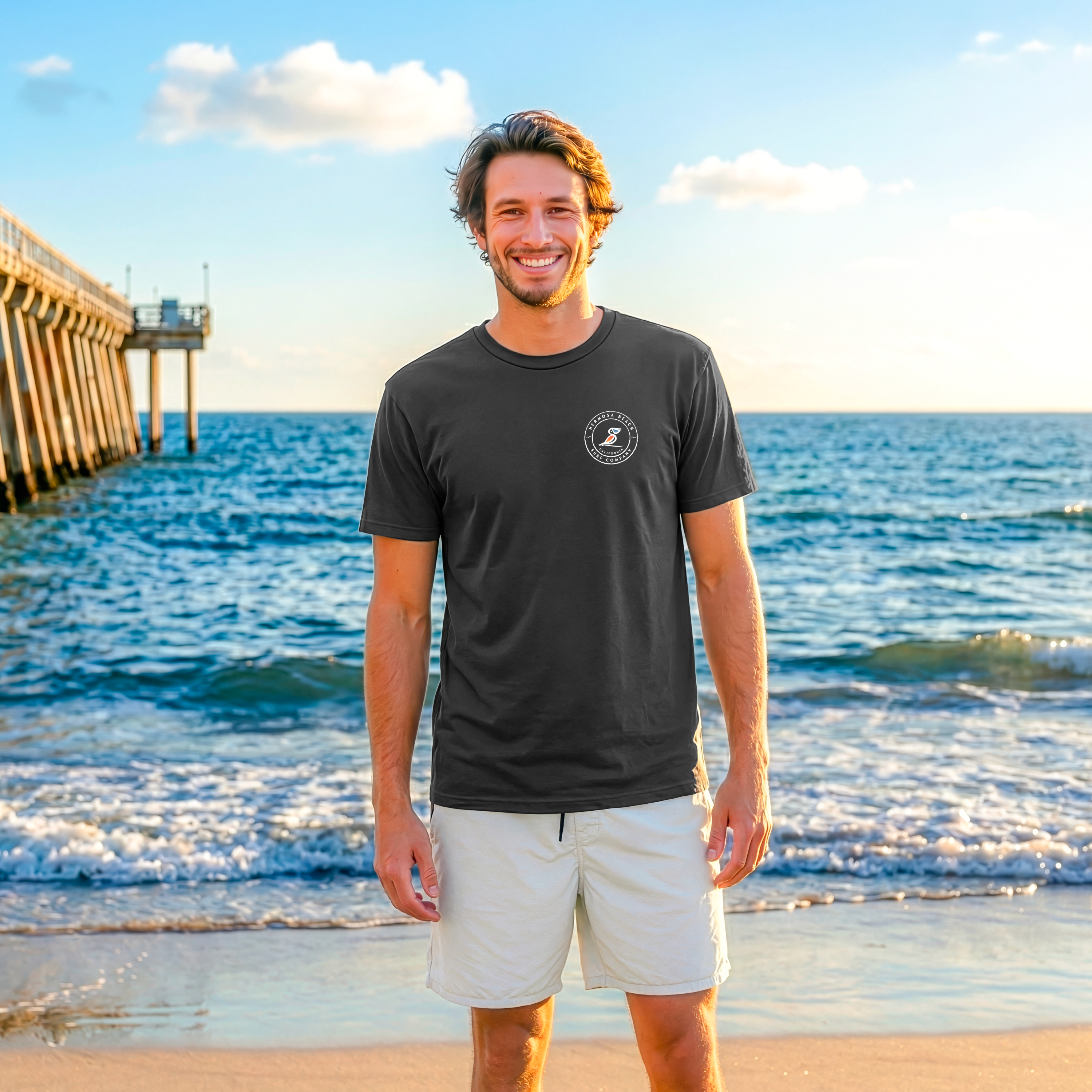 Man standing on a beach wearing a black t-shirt and light-colored shorts with a pier and ocean in the background.