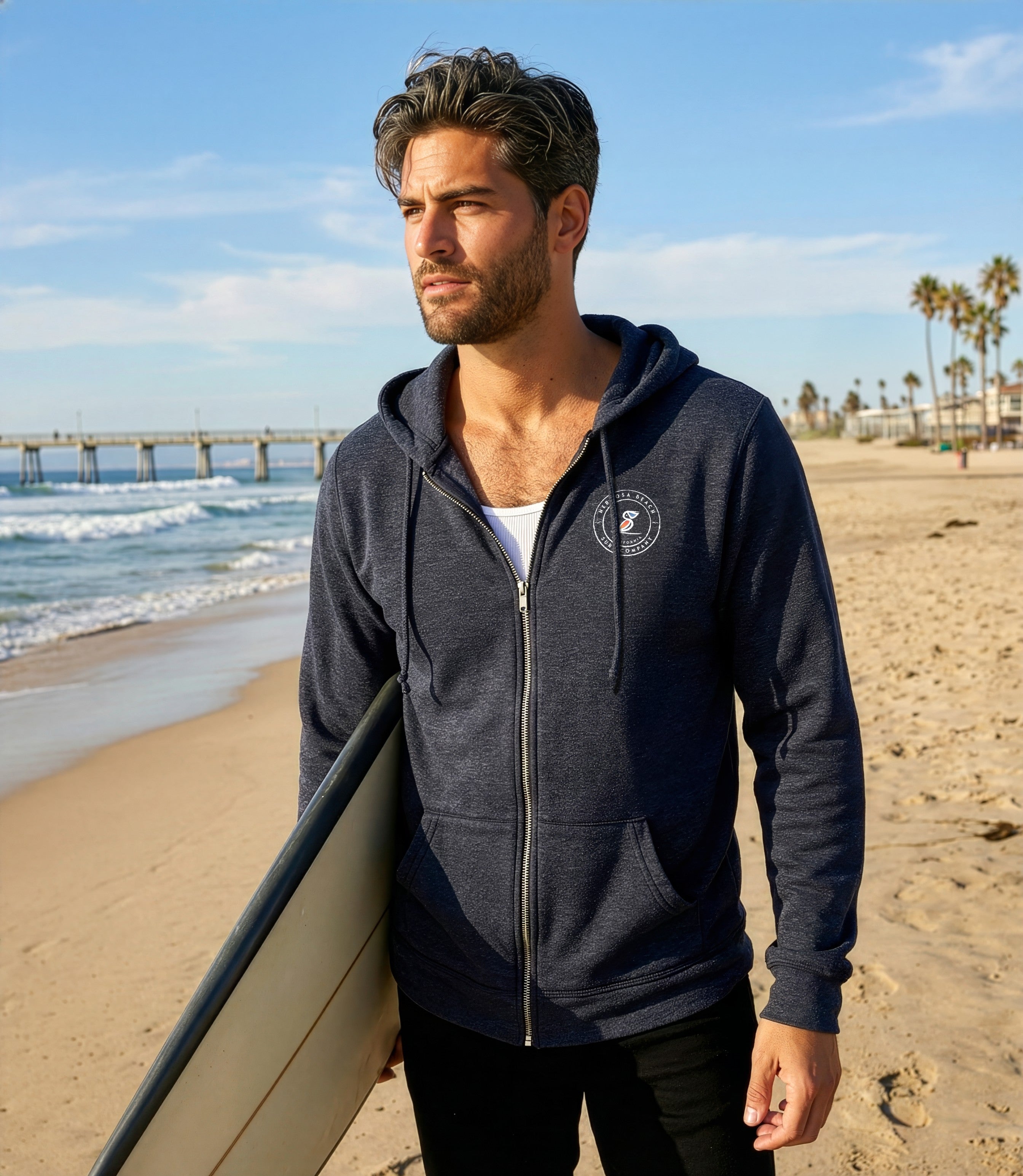 Man in a navy blue hoodie holding a surfboard on a beach with ocean and palm trees in the background