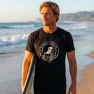 Man holding a surfboard on a beach wearing a black t-shirt with a logo.