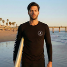 Man holding a surfboard on a beach with palm trees and a pier in the background