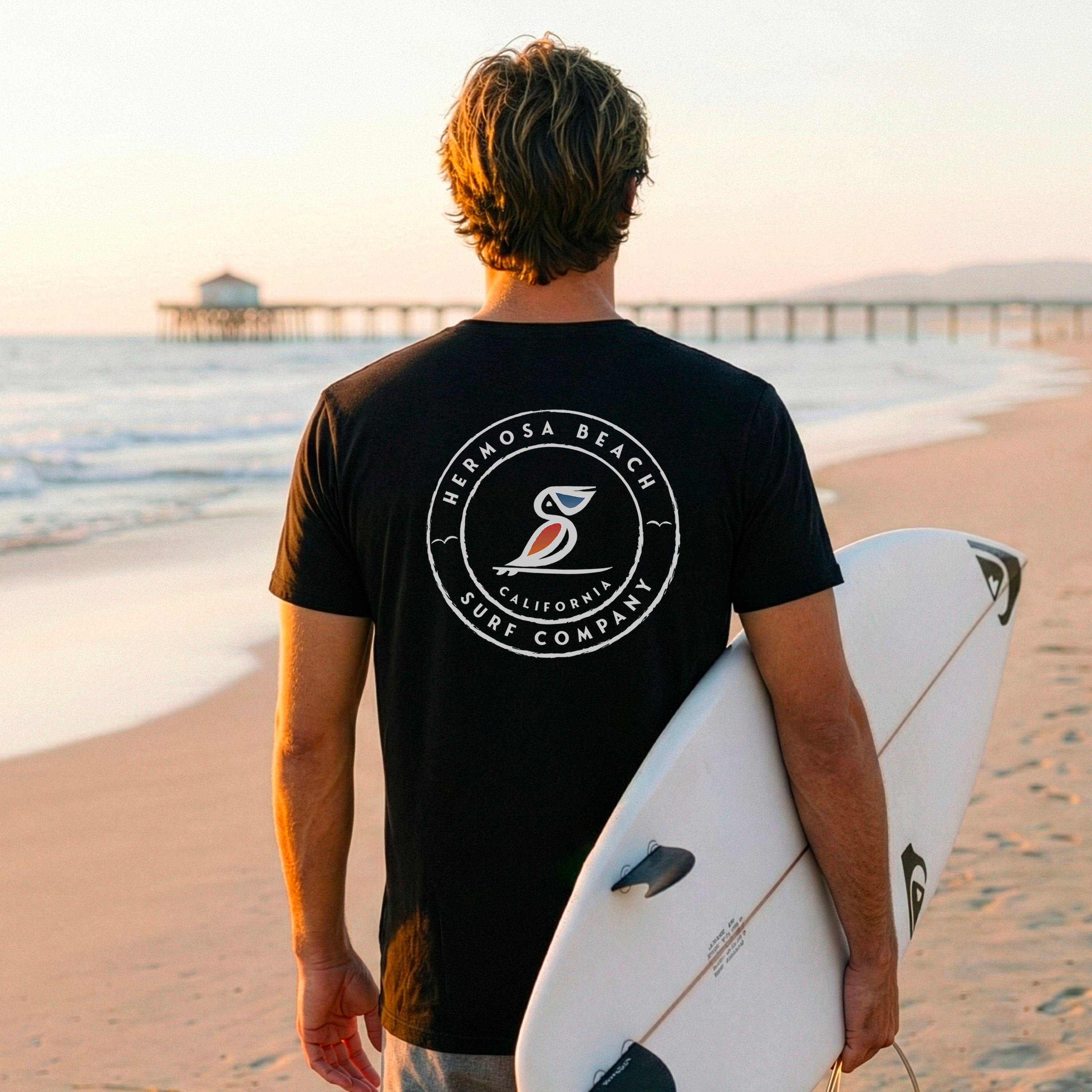 Person holding a surfboard on a beach wearing a black t-shirt with a logo.