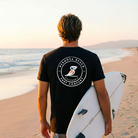 Person holding a surfboard on a beach with a sunset, wearing a black t-shirt with a logo.