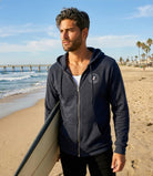 Man in a navy blue hoodie holding a surfboard on a beach with ocean and palm trees in the background