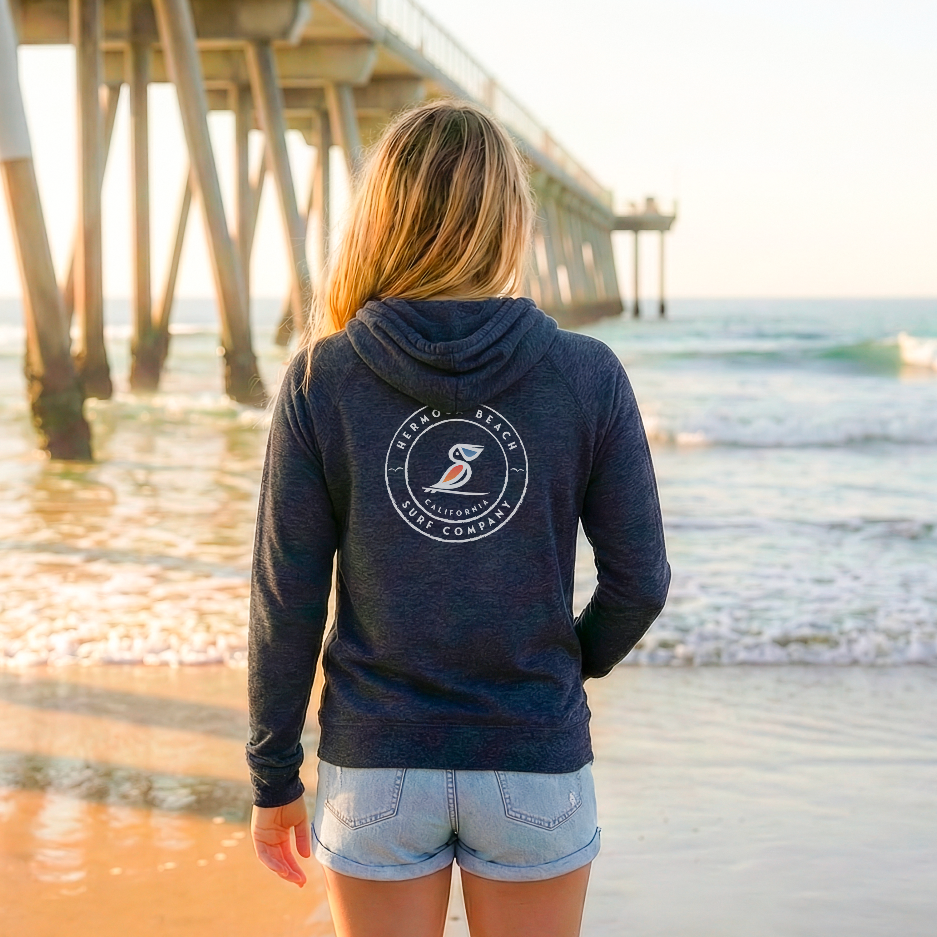 Person wearing a navy hoodie with a logo on the back, standing on a beach with a pier in the background.