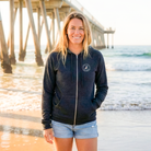 Woman wearing a navy blue hoodie with a logo on a beach with a pier in the background